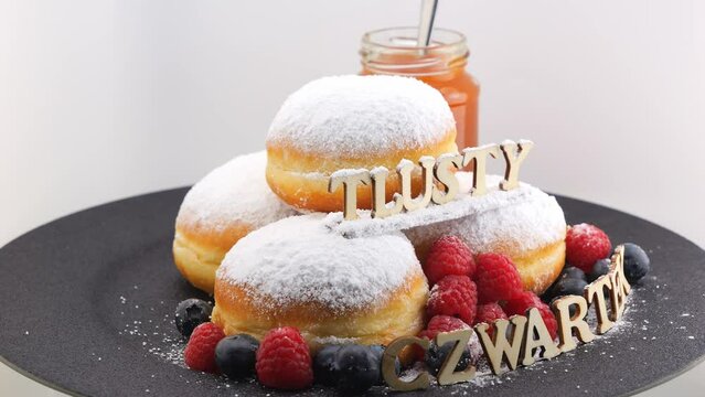 The inscription Tlusty czwartek (Fat Thursday - Polish folk-Christian holiday) in Polish. Wood letters, doughnuts with icing sugar, berries (raspberries, blueberries), jar of jam rotating on a plate. 