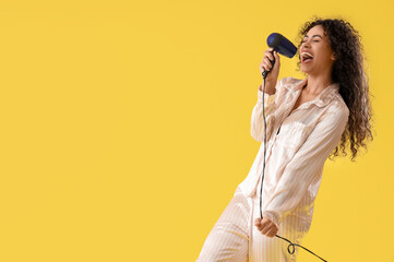 Young African-American woman with hair dryer singing on yellow background