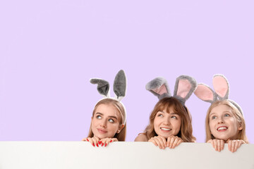 Young girls with Easter bunny ears and blank poster on lilac background