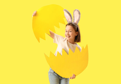 Cute little girl with Easter bunny ears and broken paper egg on yellow background