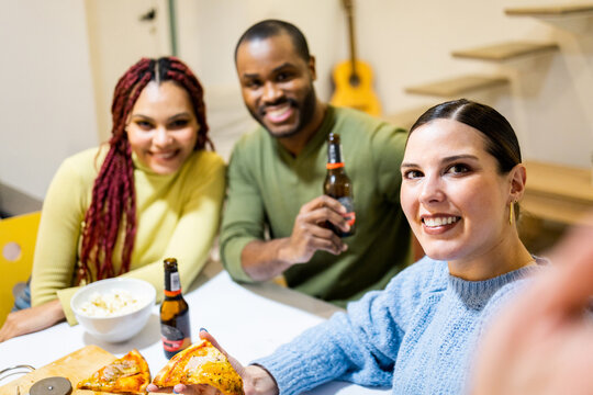 A group of 3 multi-ethnic young people gather one night for homemade pizza dinner in a charming apartment. Caucasian woman takes a photo with her mobile phone. Dinner nights among young people.