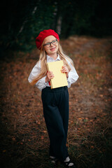 A little blonde girl in a red beret and suspenders stands with a yellow book in nature
