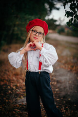 A little blonde girl in glasses, a red beret and suspenders shows a heart with her palms 