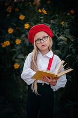 A little blonde girl in a red beret and suspenders with a book in nature