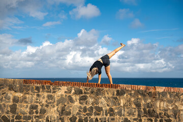 A young blonde woman does a cartwheel / hand stand on a stone wall along the sea.