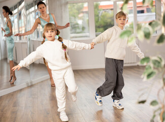 Smiling preteen girl practicing vigorous jitterbug moves with boy partner during dance lesson in choreography studio under guidance with female teacher..