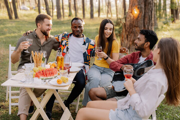 Meeting of multiracial group of friends playing guitar, singing, eating dinner and drinking wine during party in the forest