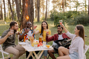 Meeting of multiracial group of friends playing guitar, singing, eating dinner and drinking wine during party in the forest