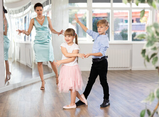 Two smiling kids, boy and girl learning cha-cha under watchful eye of female dance instructor in bright choreography studio..