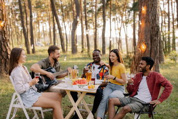 Meeting of multiracial group of friends playing guitar, singing, eating dinner and drinking wine during party in the forest
