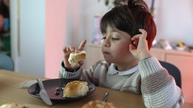 Child eating piece of bread seated at table. 5 year old boy snacking rich carb food
