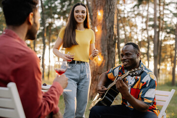 Multiracial group of friends, African man playing guitar around happy friends drinking wine