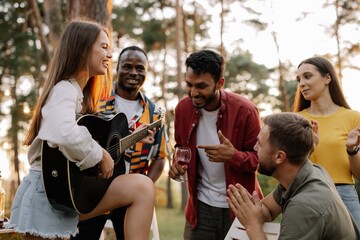 Attractive hipster woman playing guitar winning over men at dinner party with multiracial friends
