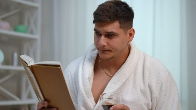 Slow motion. Close-up. A young man in a white bathrobe drinks coffee from a glass cup and reads a book while sitting in the bedroom of a modern house