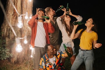 A company of multiracial friends drinking beer at party, making faces near hanging lamps