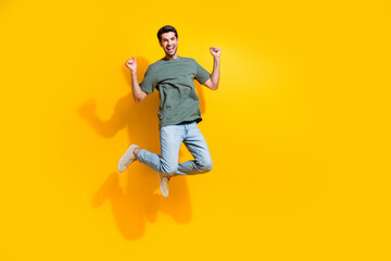 Full length photo of excited lucky guy wear khaki t-shirt jumping high rising fists empty space...