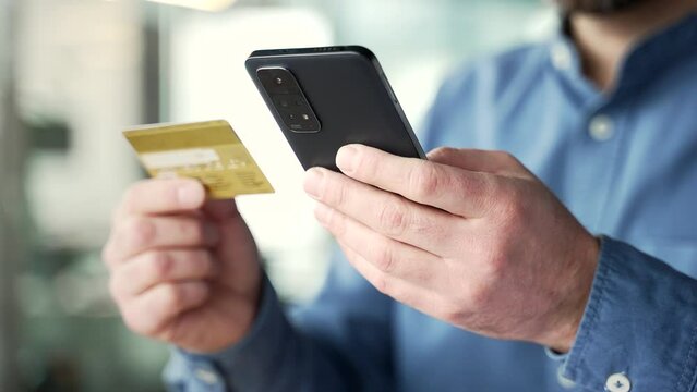 Close up of male hands holding credit card and phone. Businessman doing online shopping typing number on smartphone while sitting at workplace in business office. A man in blue shirt makes a purchase