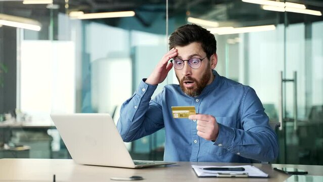 Shocked upset businessman checks his credit card on laptop while sitting at workplace in business office. Frustrated male faced fraud, money was stolen from his account. He became victim of deception