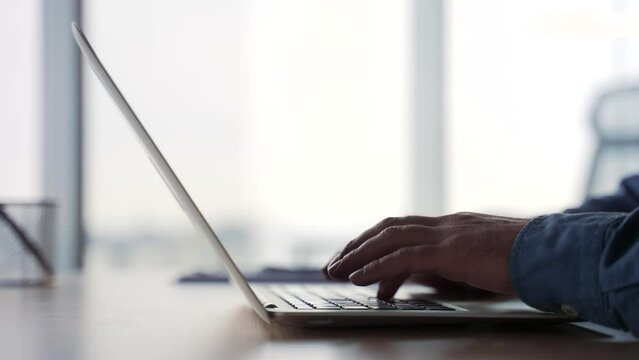 Side view. Silhouette. Close up of male hands typing on a laptop keyboard. Businessman or IT programmer uses a computer while sitting at a desk at a workplace in a business office. Man fingers texting