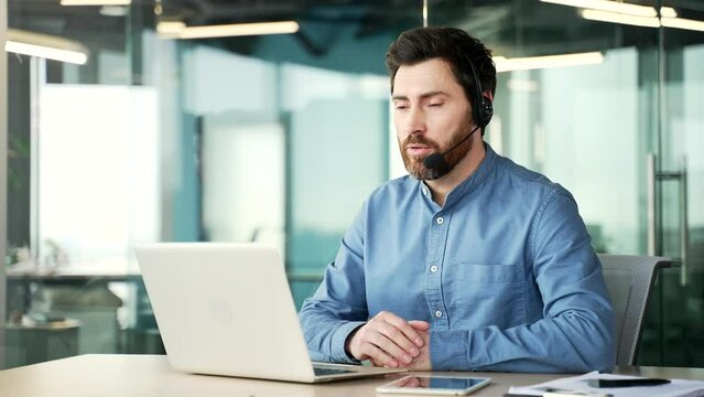 Confident handsome businessman in headset talking on a video call using laptop sitting at workplace in office. Entrepreneur has business meeting. Coach speaks remotely at an online course or training