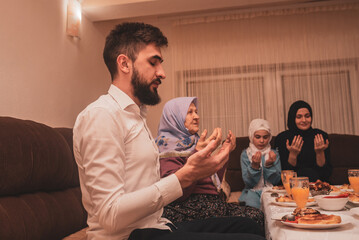 Muslim family together making iftar dua to break fasting during Ramadan dining table at home young father praying with hands up.