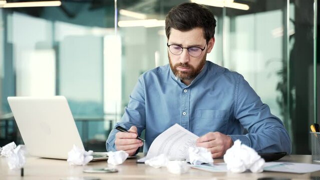 Overworked bearded financier in glasses is bored filling out documents sitting at workplace in business office. Exhausted accountant is tired of doing paper work. Man has depression, despair. Crisis