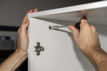 Assembling furniture at home. Tightening the bolt with a screwdriver. Cabinet installation. A man in a white T-shirt is assembling a white wardrobe. Male hands close-up.