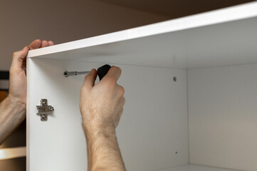 Assembling furniture at home. Tightening the bolt with a screwdriver. Cabinet installation. A man in a white T-shirt is assembling a white wardrobe. Male hands close-up.