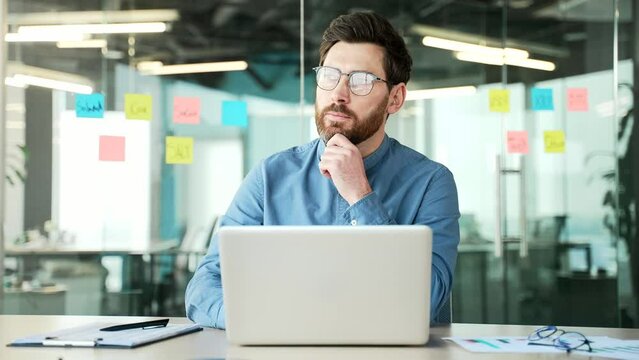 Thoughtful confident businessman works on laptop sitting at workplace in business office. Handsome entrepreneur in glasses is busy with startup project on computer or thinking about solving problem