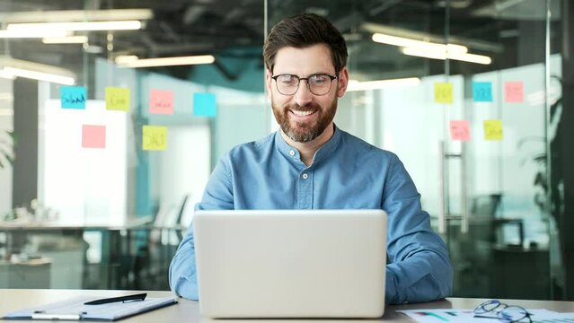 Happy manager typing on a laptop sitting at workplace in office. Smiling bearded businessman in glasses working in computer application, banking, texting a client, chatting online. busy with a project