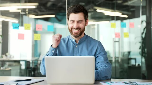 Satisfied happy handsome businessman received great news on laptop while sitting at workplace in business office. Smiling glad bearded entrepreneur reads a positive good message on the computer