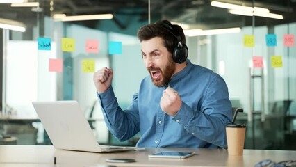 Excited businessman in wireless headphones watching sports match or competition using laptop sitting at workplace in business office. Happy man cheering for favorite team with emotional gesturing