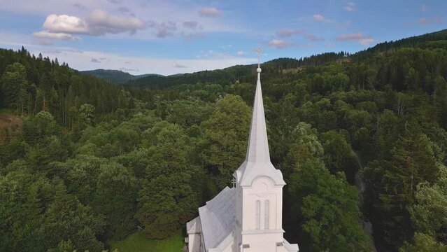 Flying by white church tower with steel cross on top in green forest nature, christian religion temple
