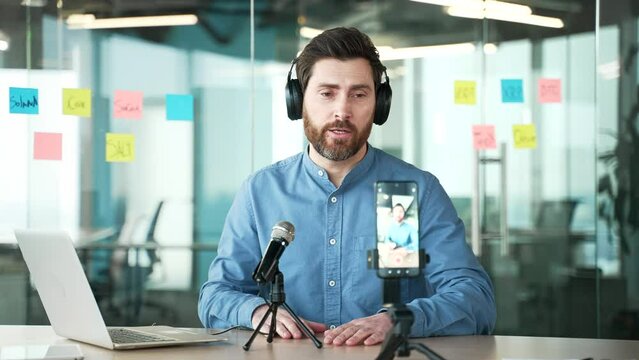 Businessman or private tutor in headphones giving video call training using smartphone microphone sitting at workplace in business office. Blogger is streaming broadcasting. Coach has an online course