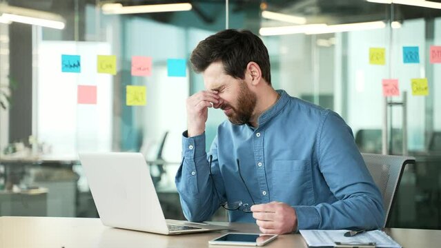 Overworked bearded businessman suffering from eye strain and headache working on a laptop sitting at workplace in business office. Tired entrepreneur takes off glasses and massages rubs bridge of nose