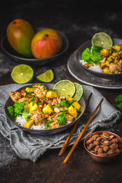 Chicken and Mango stir fry with sliced lime and coriander, in dark bowls with rice and chopsticks, mangoes in background