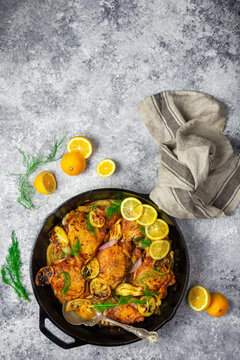 Chicken thighs in cast iron skillet with fresh and roasted lemons, fennel and shallots on gray background with sliced lemon garnish