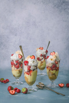 Four large parfait glasses layered with vanilla custard, cookie crumbles, whipped cream and strawberries on pale blue background