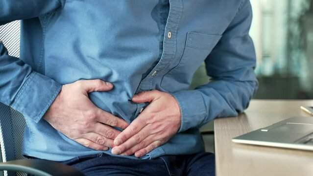 Close up. Male hands holding massaging stomach. Businessman in blue shirt feels stomach pain sitting at workplace in business office. Sick man suffers from bloating, gastritis, poisoning or bloating