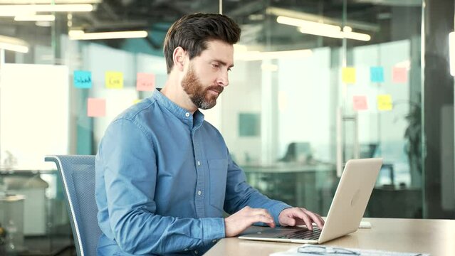 Side view. Focused businessman typing on a laptop sitting at a workplace in office. Bearded manager working in computer application, banking, texting a client, chatting online. busy with a project