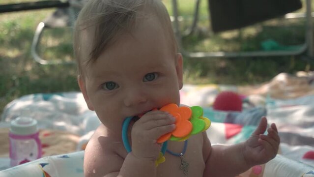 cute blue-eyed baby with a cross on his neck interestedly examines a toy rattle with his mouth, licks it, mother's hands pour warm water from the children's pool on his body
