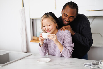 Young african american man hugging smiling asian girlfriend in sweater holding coffee in kitchen