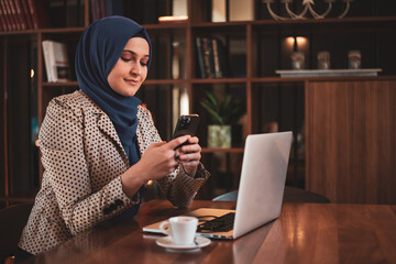 Successful smiling Arab woman in hijab working inside modern office, business woman using a smart phone Muslim woman using laptop at work