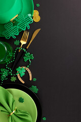 Immersed in Irish spirit: Crafting your St. Paddy's table tale. Top view vertical shot of plates, cutlery, green hat, green beer, party eyewear, festive decor on black background with greeting zone