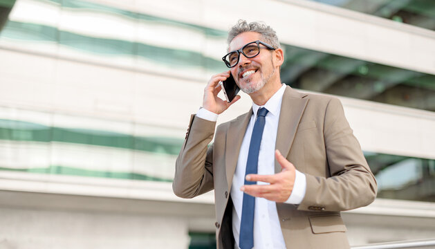 Mature businessman laughing during a phone call, in a modern urban landscape. His relaxed posture and genuine smile convey a successful professional enjoying a moment of lighthearted communication