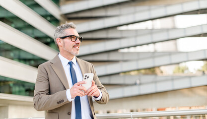 Cheerful grey-haired businessman engaging with his smartphone outdoors, with a modern office...