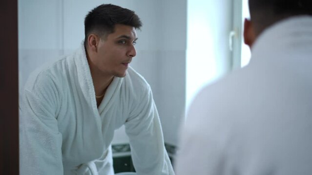 Slow motion. A young man in a white bathrobe carefully examines himself in the mirror on the bathroom wall. A man washes his face with water while standing in the bathroom