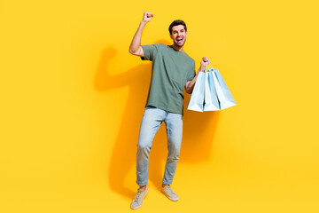 Full length photo of positive lucky guy wear khaki t-shirt rising fist holding shoppers isolated yellow color background