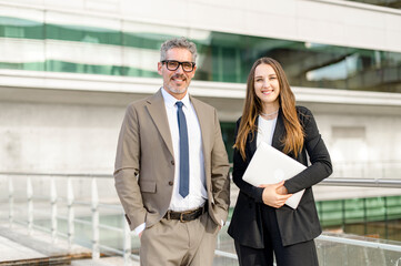 Fototapeta premium Standing side by side, the seasoned businessman and his young female colleague share a professional moment, woman holding laptops and both dressed in business attire against a modern building backdrop