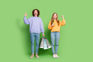 Fototapeta premium Full length photo of two delighted people raise fists hold shop bags use smart phone isolated on green color background